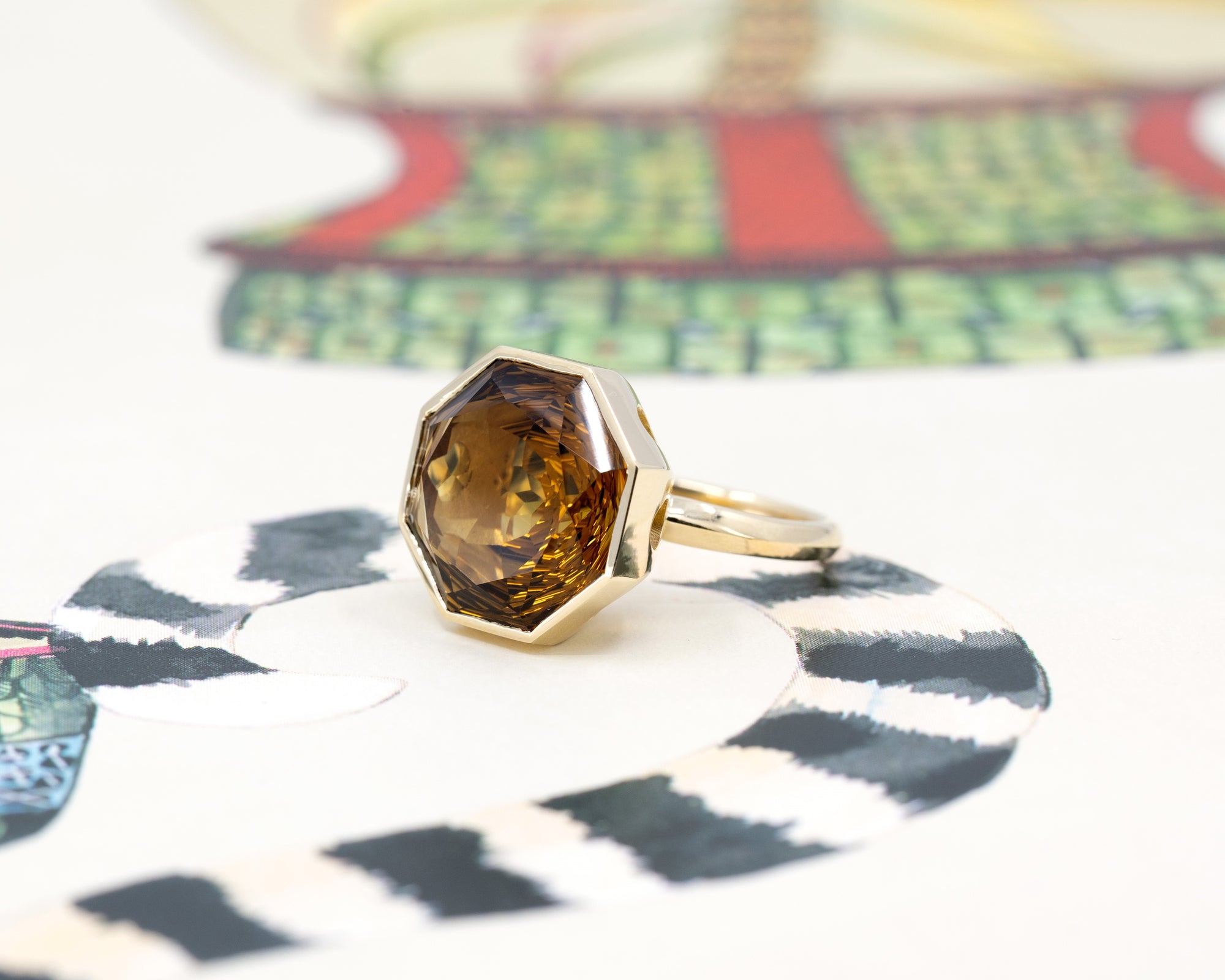 Octagonal dark orange citrine ring in yellow gold, glowing against a vibrant color backdrop, designed by Bena Jewelry.