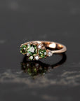 Unique rose gold ring with green gemstones, including sapphire and garnet, shown against a dark background. Made by Bena Jewelry, Montreal, Canada.
