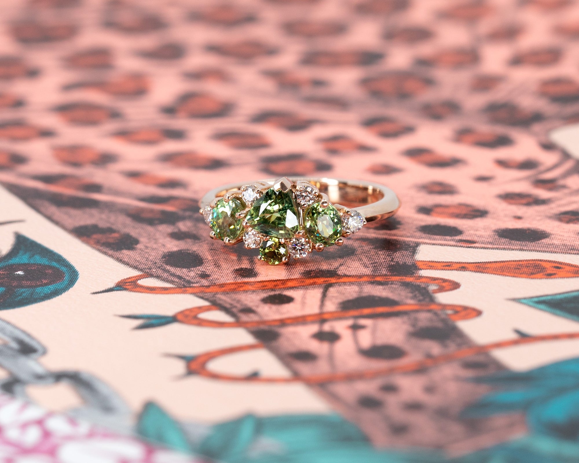 Custom designer ring with green gemstones in rose gold, shown against a soft-colored backdrop. Created by Bena Jewelry, Montreal, Canada.