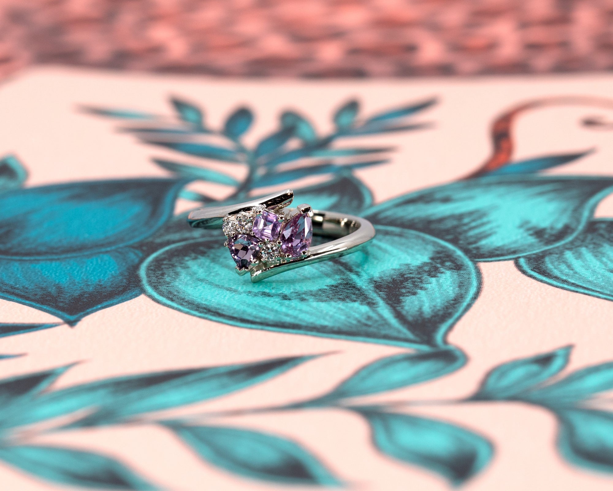 White gold Kink Avalanche ring by Bena Jewelry, featuring vivid purple and pink sapphires in trillion, pear, and round diamond cuts, with a dynamic twist band design, displayed against a vibrant, colorful background.