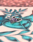 White gold Kink Avalanche ring by Bena Jewelry, featuring vivid purple and pink sapphires in trillion, pear, and round diamond cuts, with a dynamic twist band design, displayed against a vibrant, colorful background.