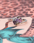 Side view of the Kink Avalanche white gold ring's dynamic twist band design, featuring purple and pink sapphires and diamonds, on a colorful background.