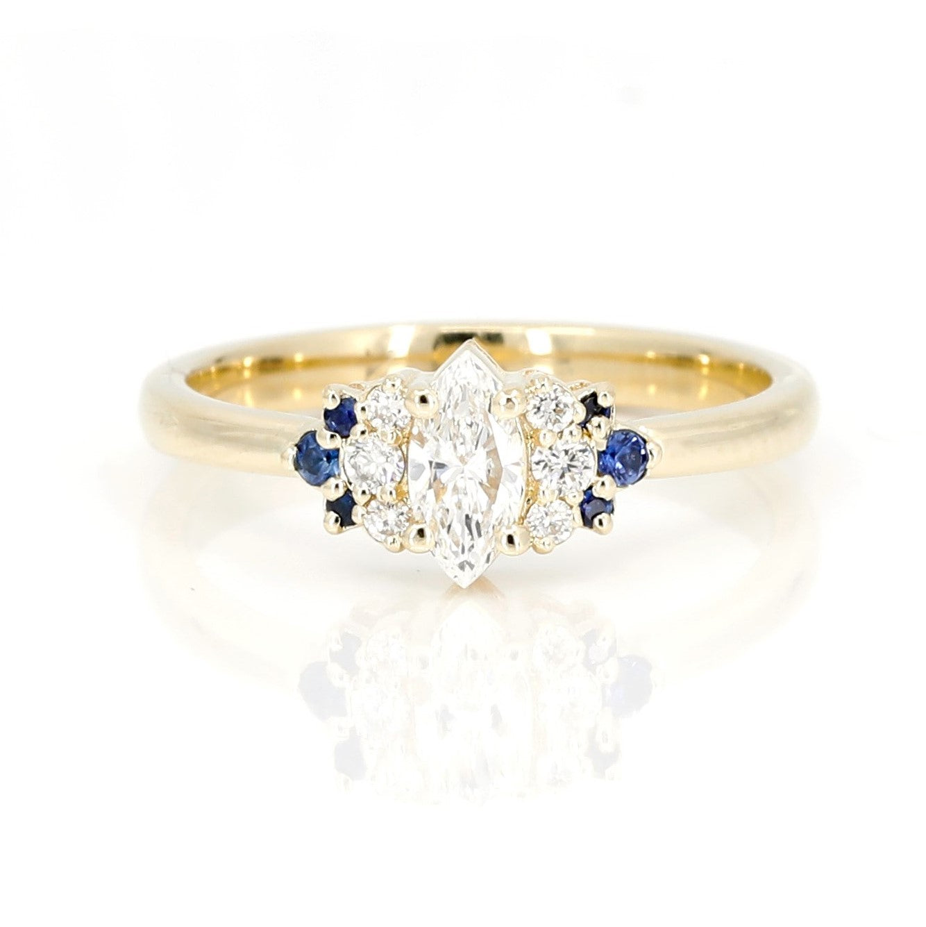 Yellow gold engagement ring featuring a marquise-cut diamond, round diamond accents, and royal blue sapphires by Bena Jewelry, photographed on a white background.