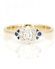 Yellow gold engagement ring featuring a marquise-cut diamond, round diamond accents, and royal blue sapphires by Bena Jewelry, photographed on a white background.
