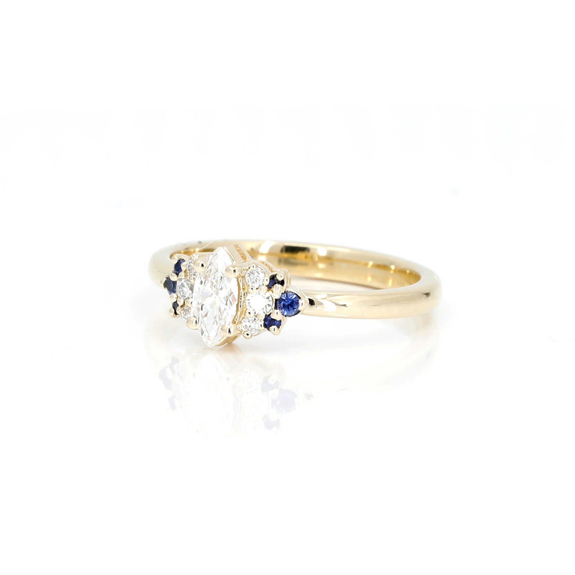 Yellow gold engagement ring with a central marquise-cut diamond, flanked by sparkling diamonds and royal blue sapphires, handcrafted by Bena Jewelry, displayed on a white background.