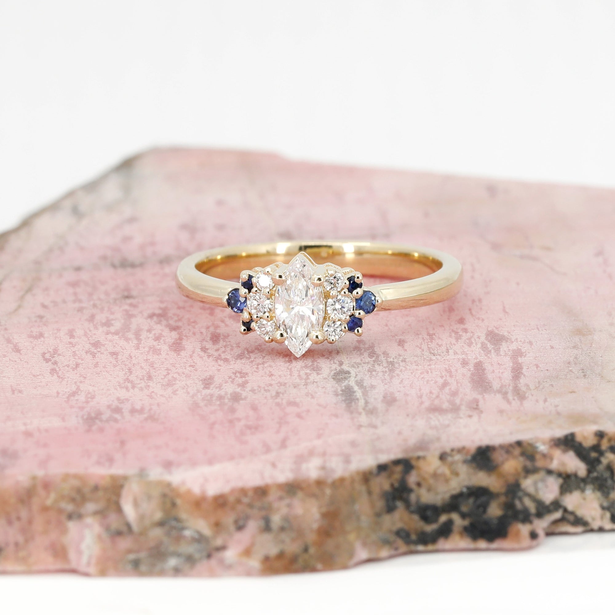 Custom bridal ring in yellow gold with a marquise diamond, diamonds, and sapphires, crafted by Bena Jewelry, displayed on a pinkish background.