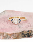 Custom bridal ring in yellow gold with a marquise diamond, diamonds, and sapphires, crafted by Bena Jewelry, displayed on a pinkish background.
