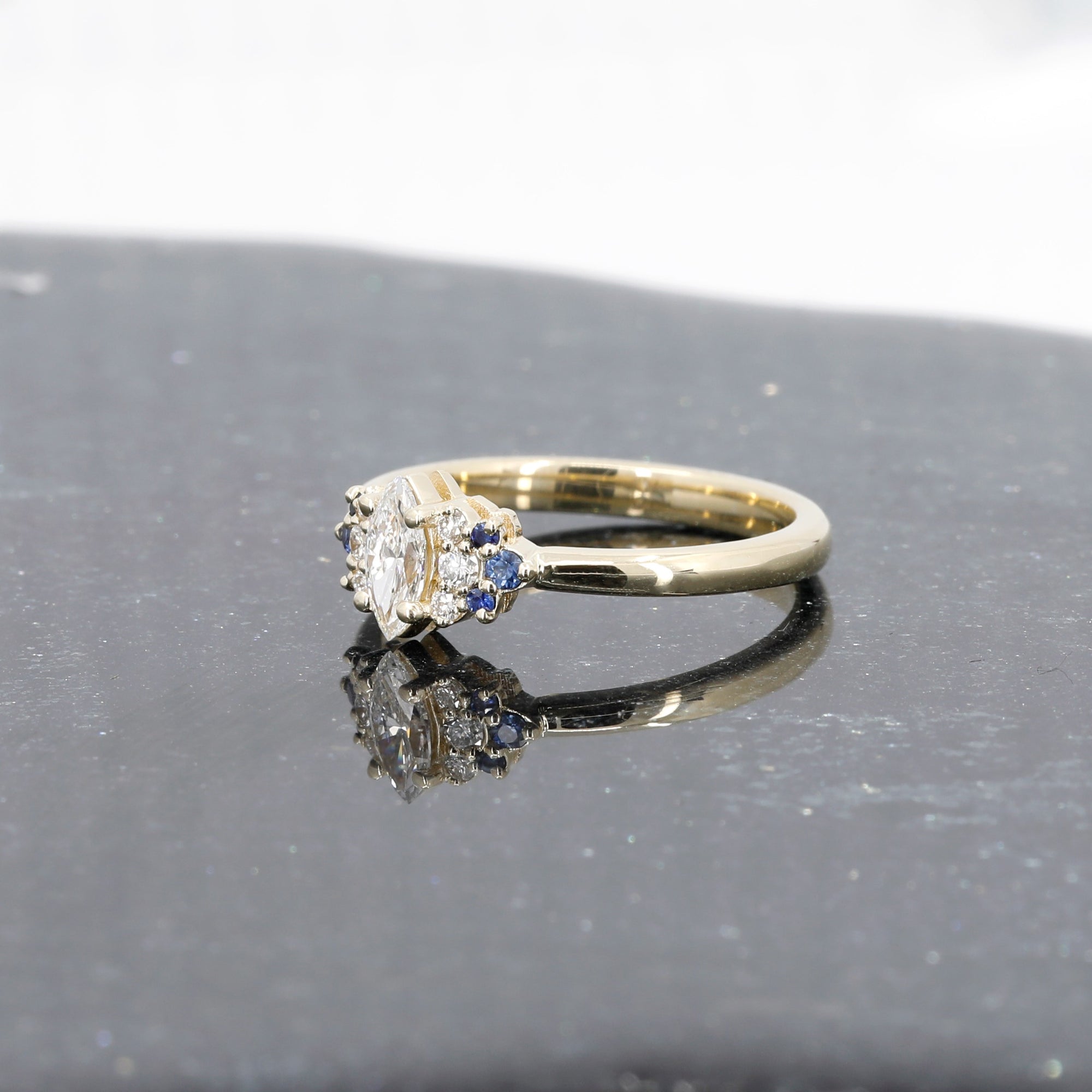Elegant marquise diamond and sapphire ring in yellow gold by Bena Jewelry, photographed on a dark glossy background, fine bridal jewelry handmade in Montreal.
