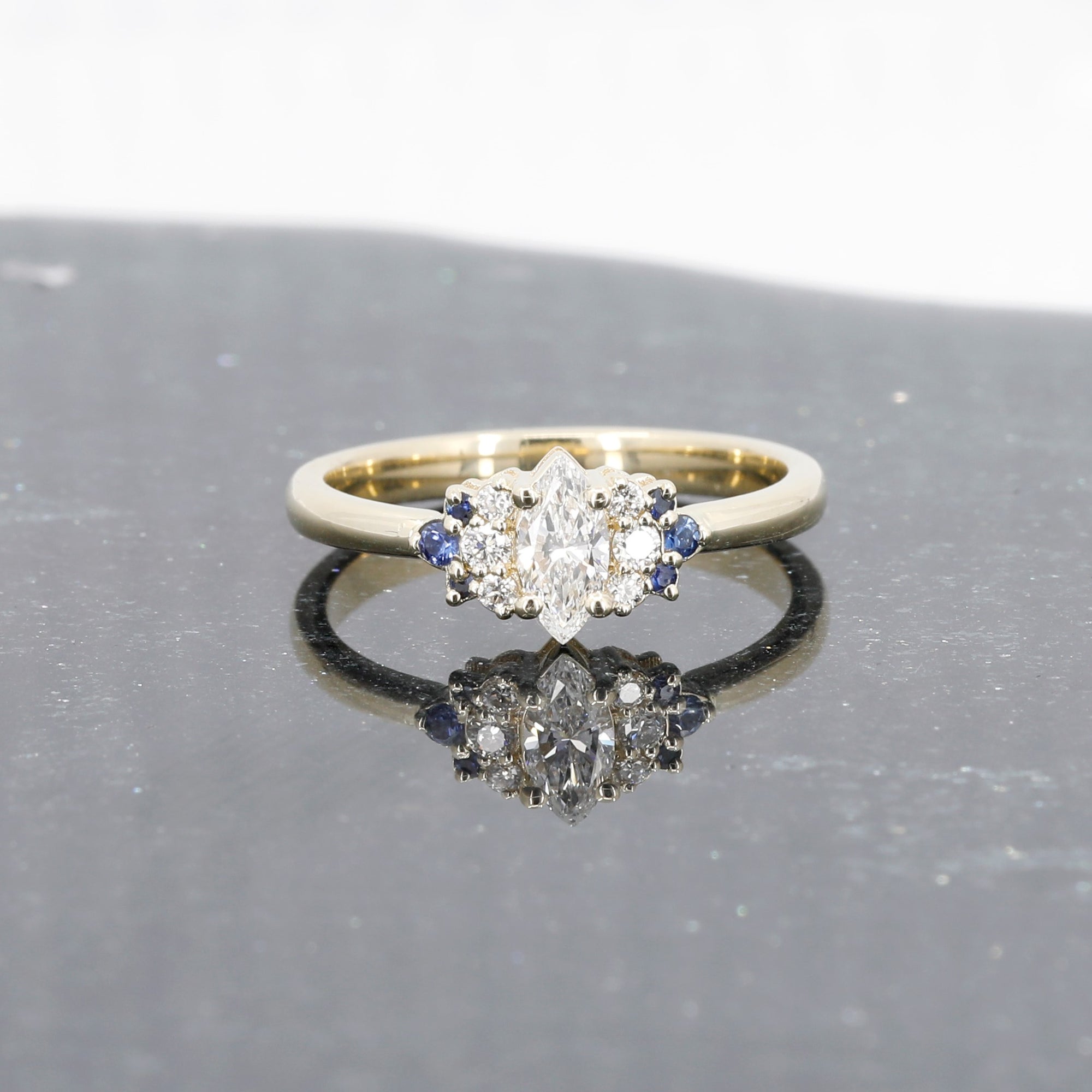 Yellow gold ring featuring a marquise diamond, diamond accents, and royal blue sapphires, designed by Bena Jewelry, displayed on a black matte background.