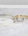 Yellow gold ring featuring a marquise diamond, diamond accents, and royal blue sapphires, designed by Bena Jewelry, displayed on a black matte background.