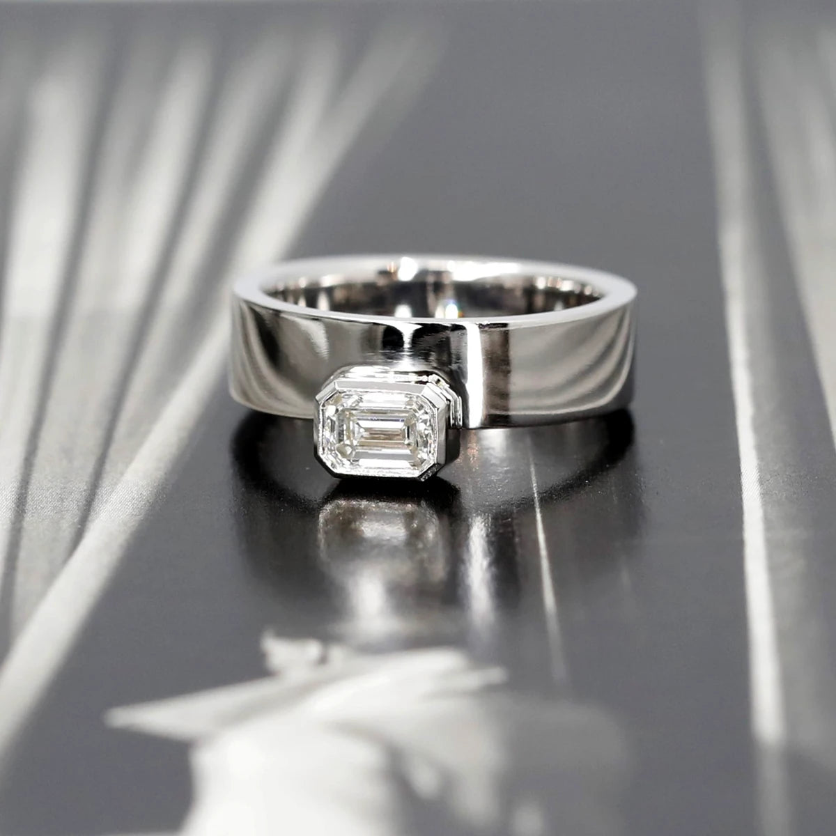 Edgy white gold ring with an emerald-cut diamond in a bezel setting, captured in a moody black-and-white photo. The ring is in sharp focus, while a woman in an overcoat appears blurred in the background, enhancing the ring’s bold statement.
