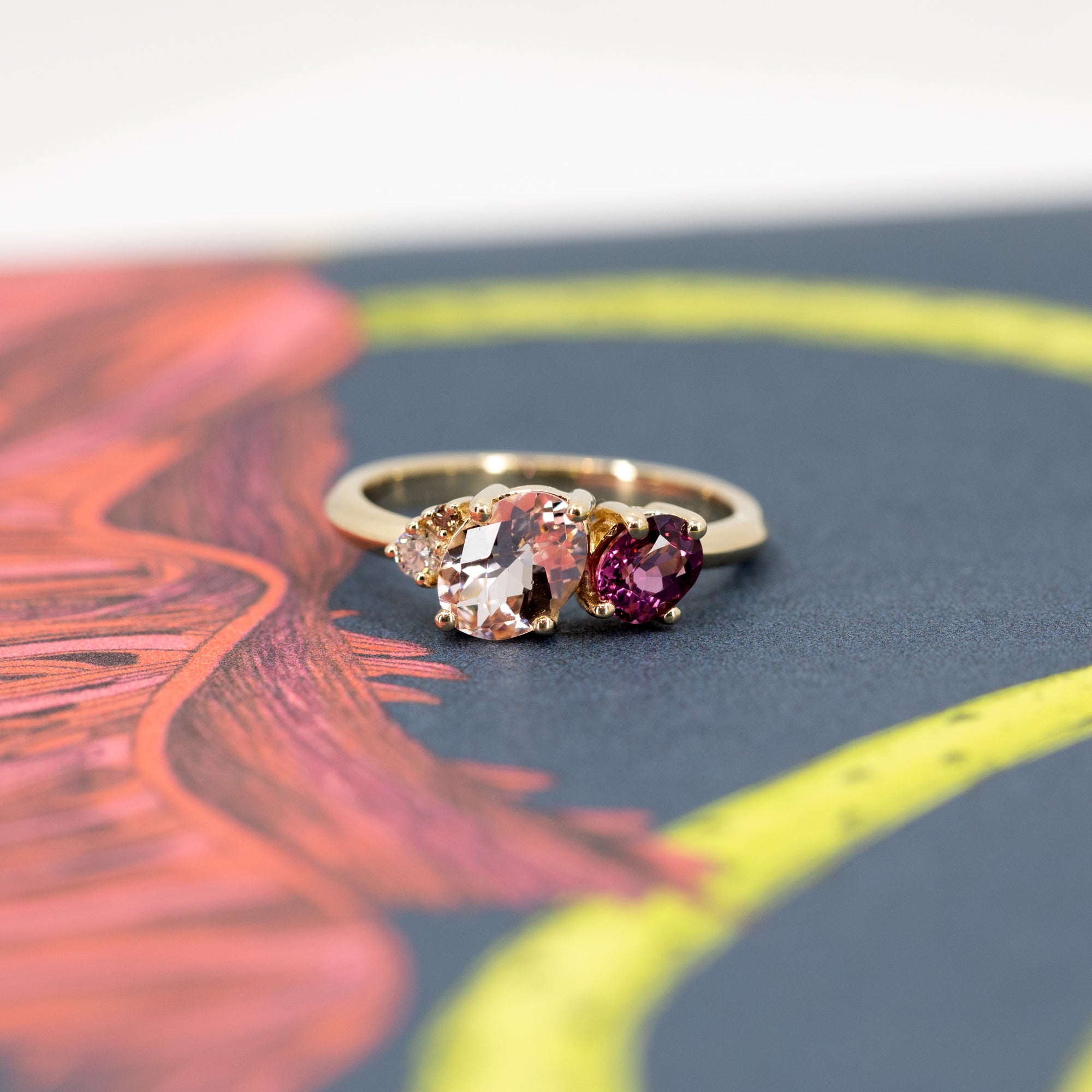 Trio ring in yellow gold with morganite, rhodolite garnet, and diamond, displayed on a warm-toned background. Designed by Bena Jewelry, handmade in Montreal, Canada.