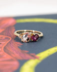 Trio ring in yellow gold with morganite, rhodolite garnet, and diamond, displayed on a warm-toned background. Designed by Bena Jewelry, handmade in Montreal, Canada.