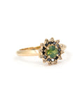 Close-up of a handmade green sapphire ring by Bena Jewelry, with black and brown diamond halo and faceted yellow gold band, photographed on white surface.