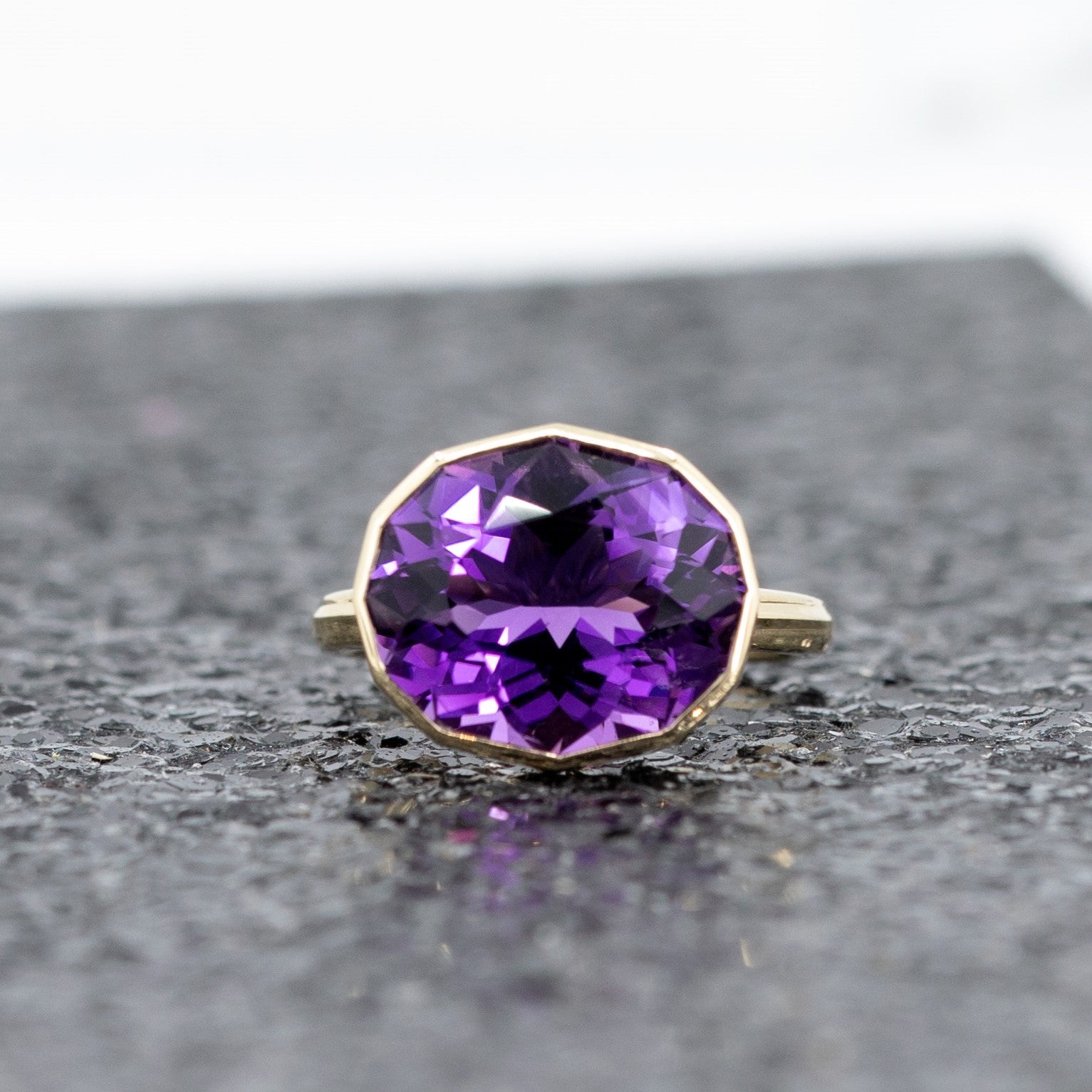 Front viewof a statement oval octagonal amethyst ring in yellow gold, glowing against a dark glittery background.