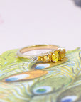 Yellow gold engagement ring featuring a vivid yellow oval sapphire center stone, flanked by orange princess cut sapphires and diamonds, displayed against a floral background.