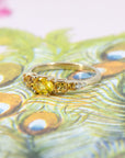 Close-up of the yellow gold engagement ring highlighting the vivid yellow oval sapphire center stone, orange princess cut sapphires, and diamond accents, set against a floral backdrop.