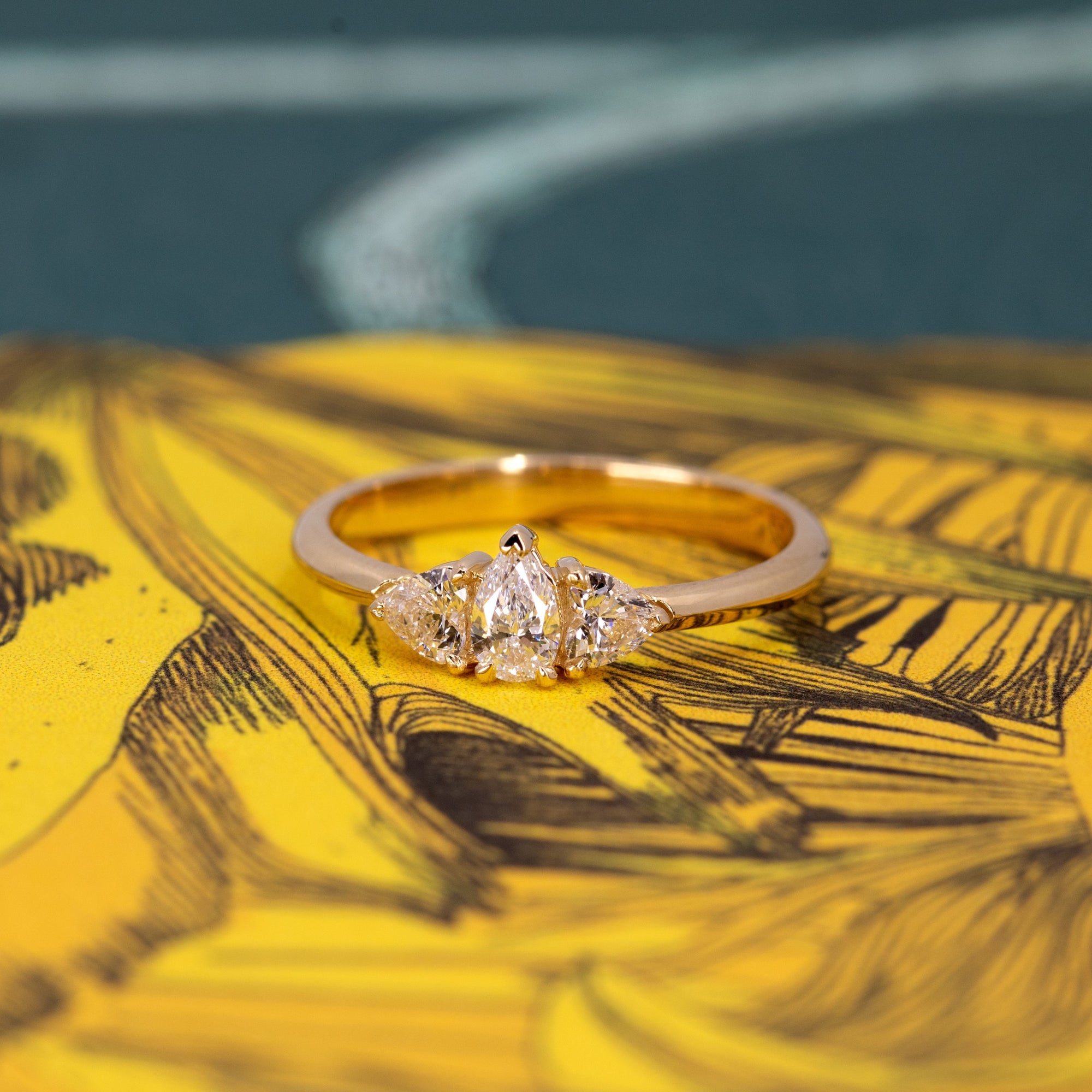 The Heart & Pear Trinity Diamond Ring, a timeless bridal ring displayed against a colored backdrop.