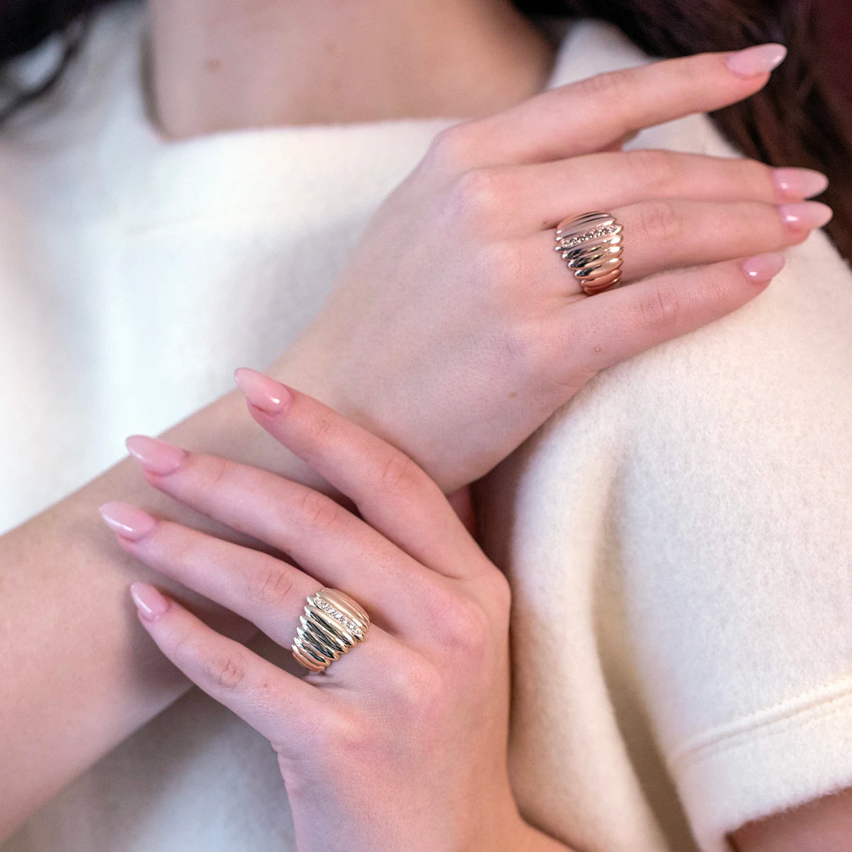 A close-up image showcasing two hands wearing bold, sculptural rings with layered designs. One ring features a mix of gold and diamond accents, adding elegance and texture. The soft, neutral-toned outfit complements the refined and modern jewelry style.