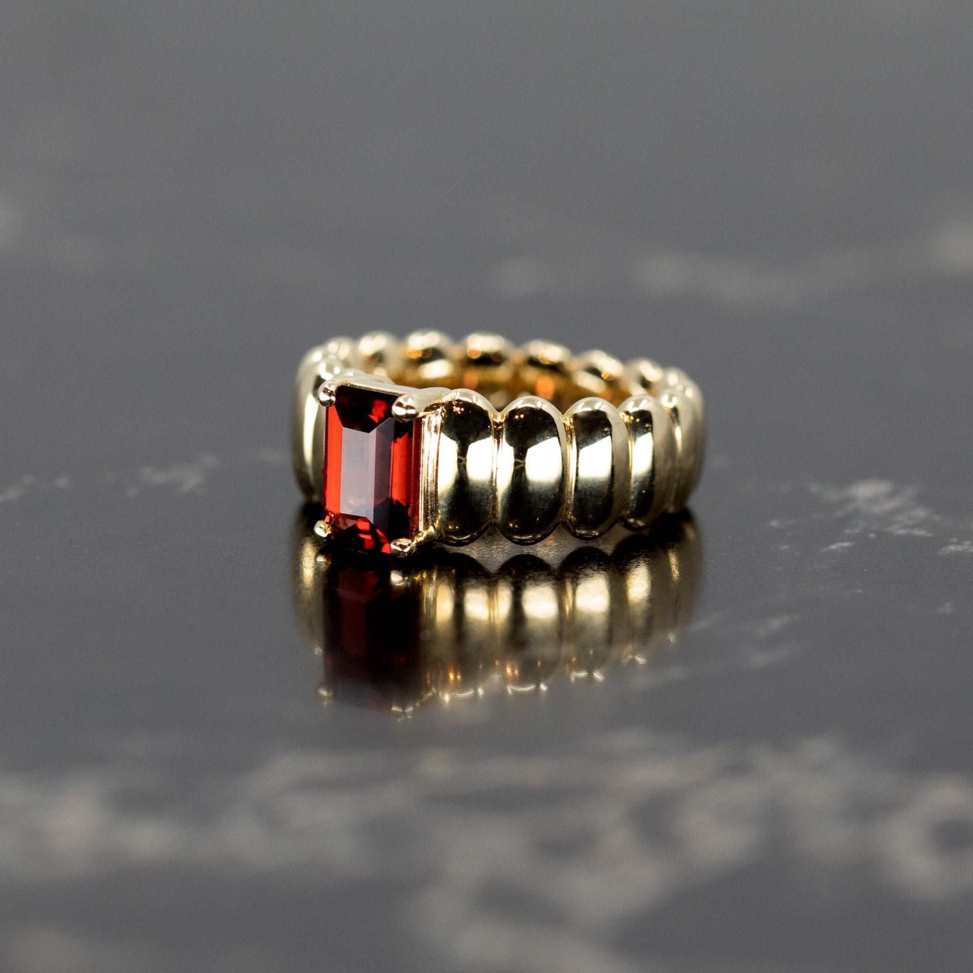 The Pyrope Pigalle Ring placed on a dark, textured surface, highlighting the yellow gold’s warm glow and the richness of the pyrope garnet, designed by Bena Jewelry and crafted in Montreal, Canada.