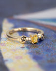 Artistic composition of a yellow gold ring with an emerald-cut yellow diamond and brown side diamonds, showcased on a soft-toned colored background.