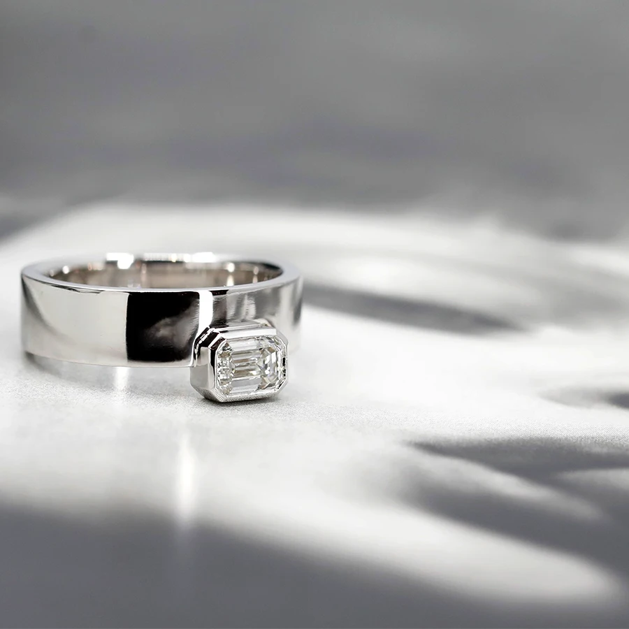 White gold ring with an emerald-cut diamond in a closed setting, captured on a mysterious black-and-white image of a woman's partially obscured face. The ring’s bold and boxy design by Bena Jewelry makes a striking statement, blending elegance with edgy allure. Designed by the Canadian independent jewelry brand, available at Ruby Mardi.