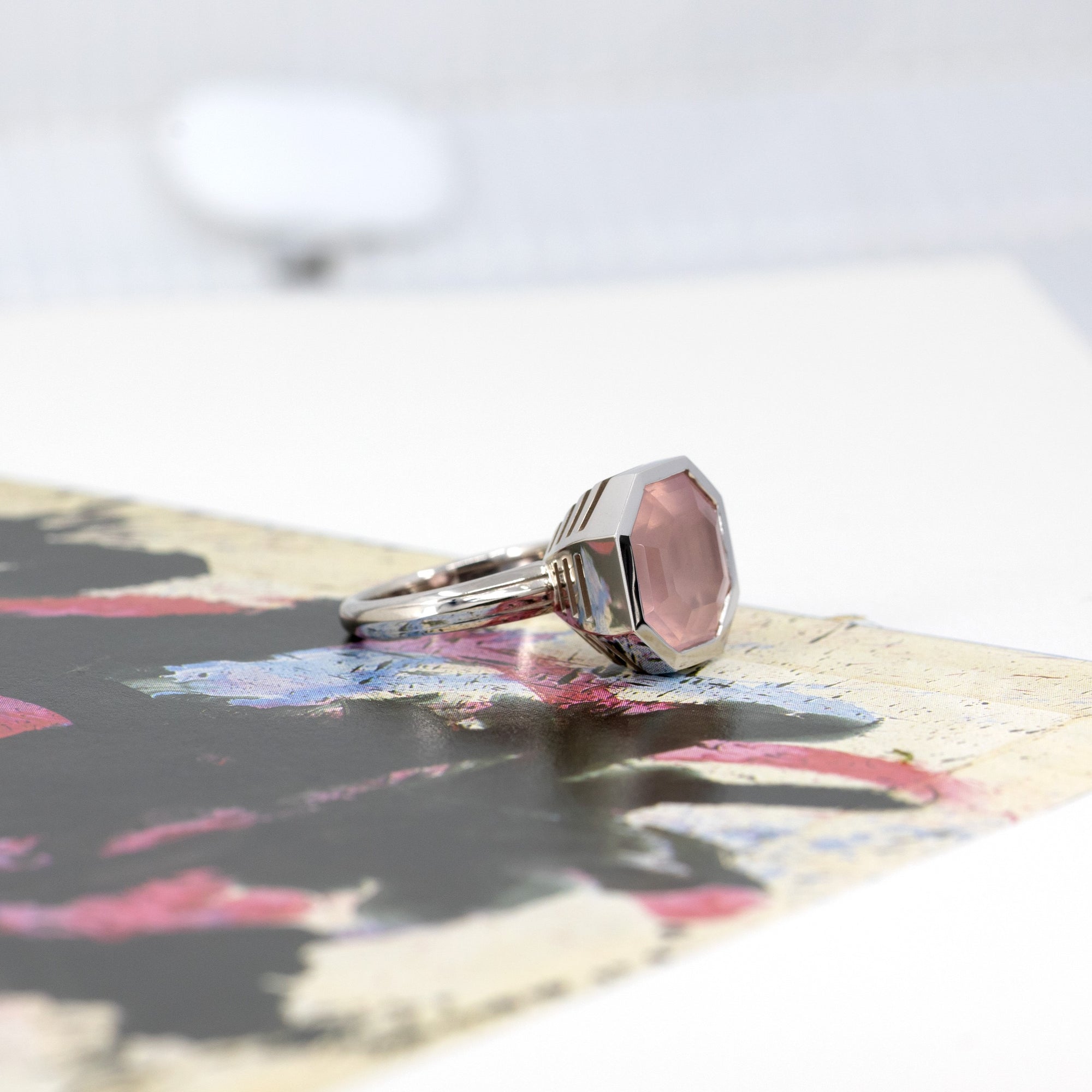 Pink rose quartz bezel setting ring in white gold, glowing against a colored backdrop, designed by Bena Jewelry.