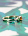 Elegant yellow gold ring with an orange sapphire and green garnet accents, displayed on a soft-toned background. Designed by Bena Jewelry in Montreal.