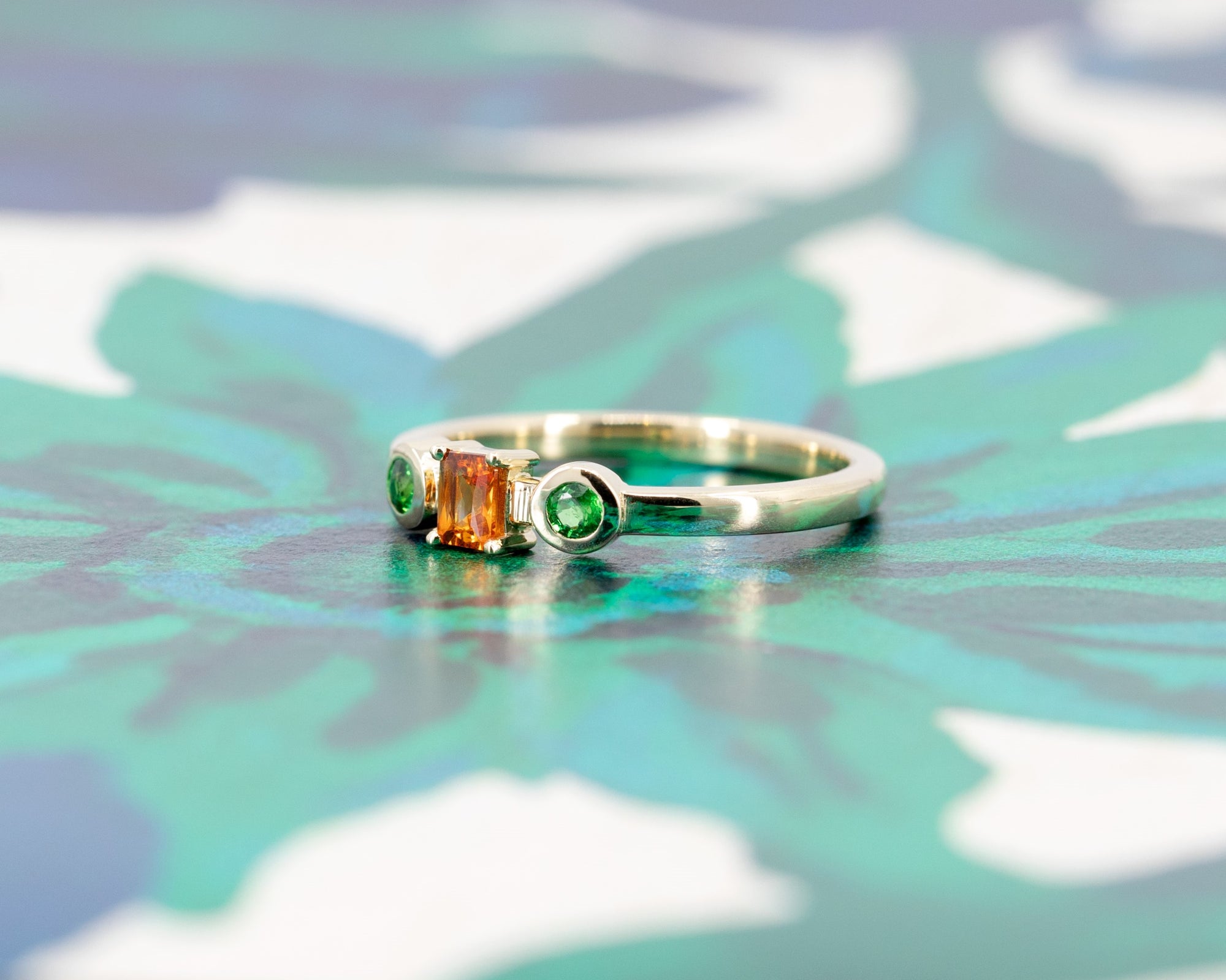 Colorful gemstone ring in yellow gold with a baguette-cut orange sapphire and green garnets, styled on a pastel backdrop by Bena Jewelry.