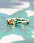 Colorful gemstone ring in yellow gold with a baguette-cut orange sapphire and green garnets, styled on a pastel backdrop by Bena Jewelry.