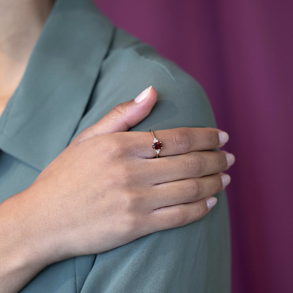 Close-up of a woman’s hand on her shoulder wearing the Désir Pyrope Garnet Ring in yellow gold featuring a vibrant oval garnet and diamonds handmade by Bena Jewelry in Canada
