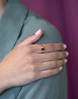 Close-up of a woman’s hand on her shoulder wearing the Désir Pyrope Garnet Ring in yellow gold featuring a vibrant oval garnet and diamonds handmade by Bena Jewelry in Canada