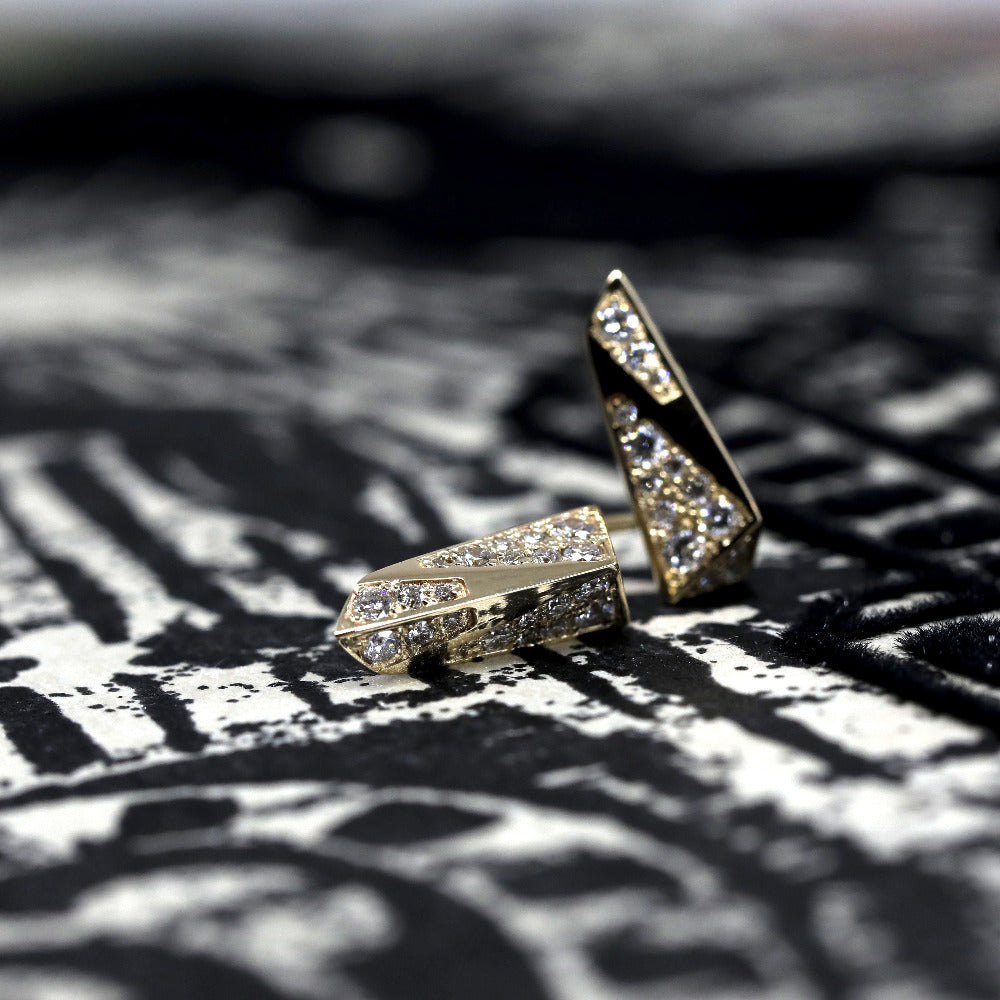 A photography of a fashion and modern pair of gold diamond stud earrings taken on a black and white textured background from Jean-Paul Gaultier. These earrings show a lot of power and energy. These unisex studs are an original design from designer Bena Jewelry.