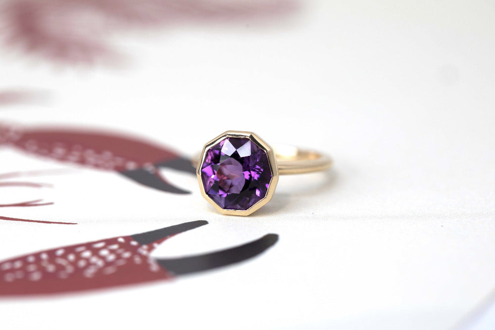 Contemporary yellow gold ring with a deep violet round amethyst, part of the Fancy Edgy collection, displayed on a white and red backdrop.