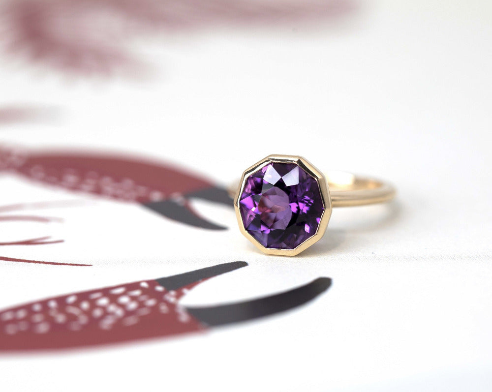 Round amethyst statement ring in yellow gold, designed by Canadian jeweler Bena Jewelry, photographed on a two-tone white and red surface.