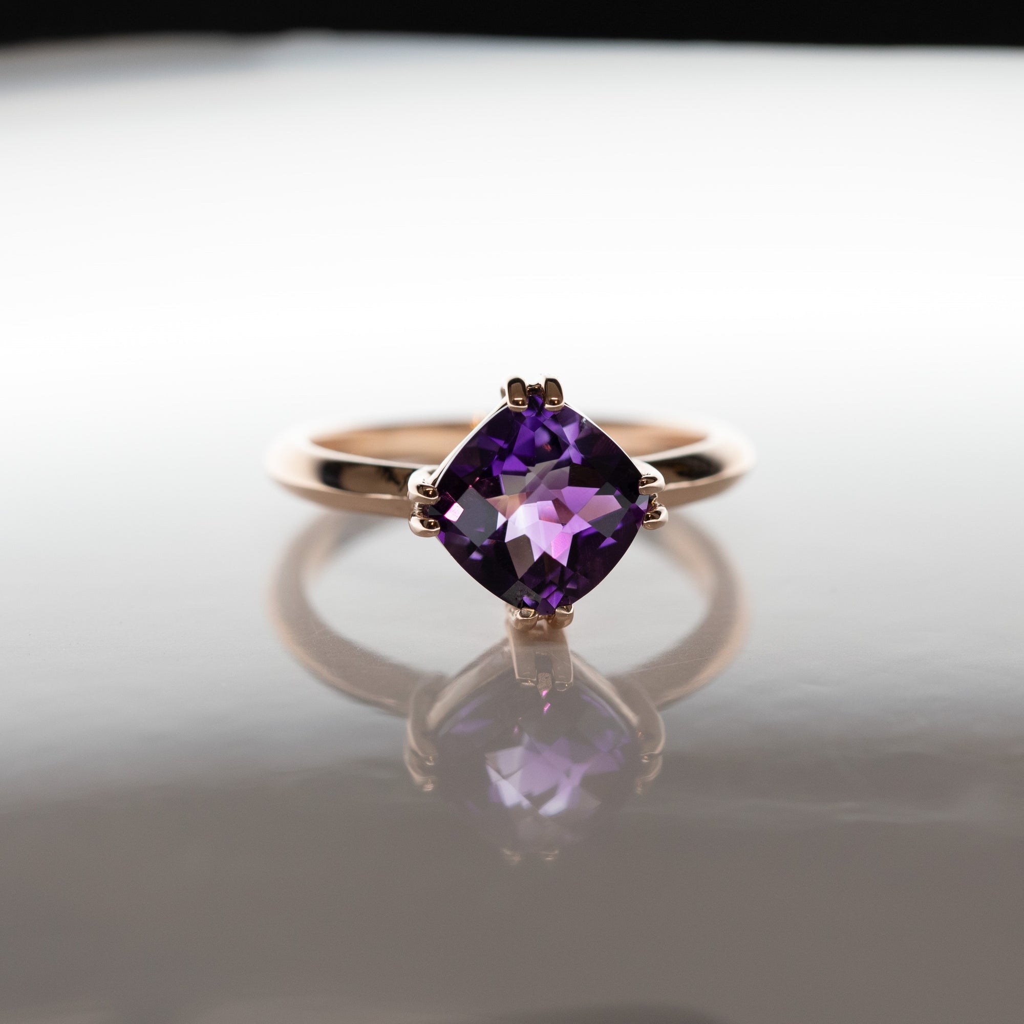 The cushion-cut amethyst ring reflects beautifully on a mirrored surface, showcasing its vibrant purple gemstone and sleek rose gold design.
