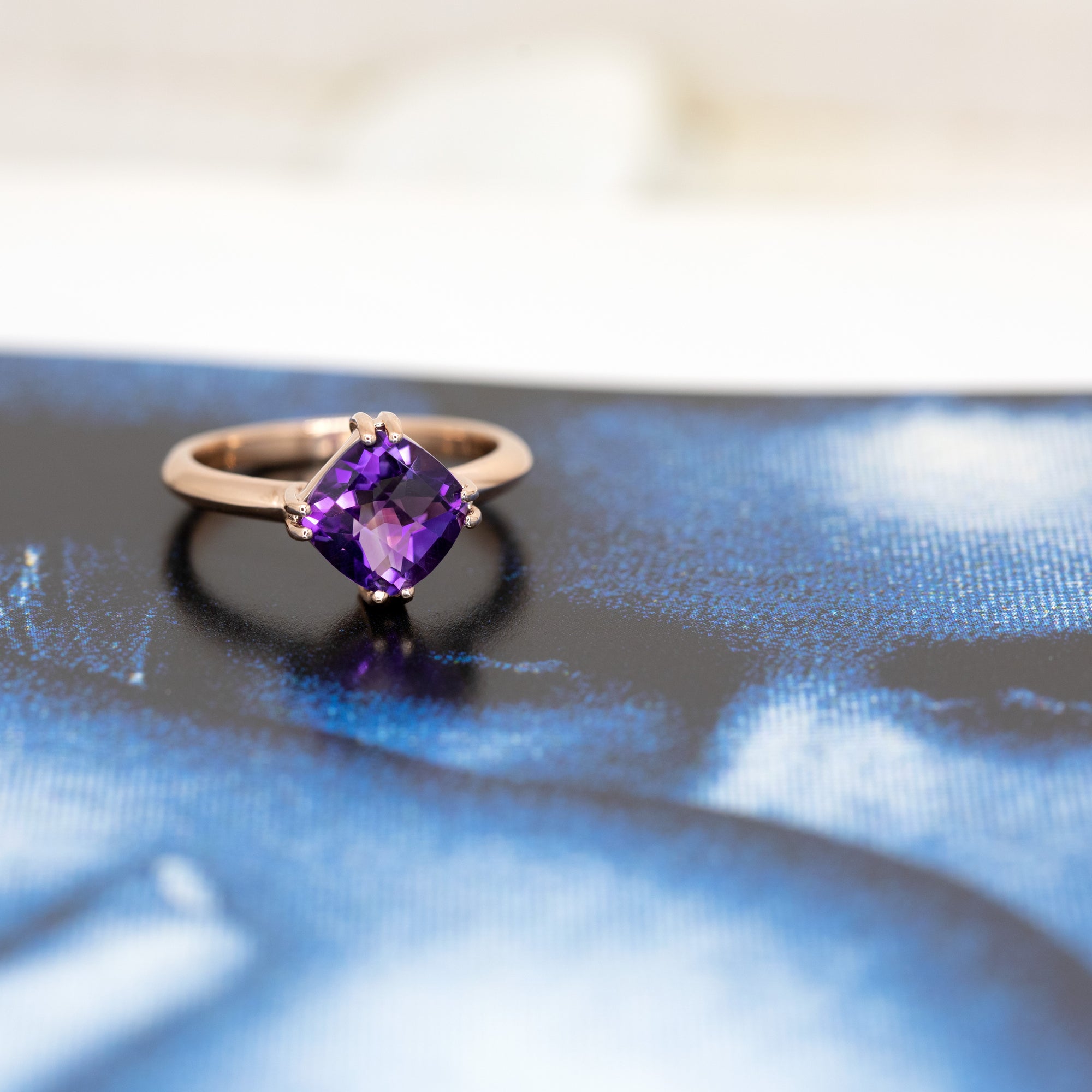 A bold cushion-cut amethyst ring glows against a dark background, emphasizing its deep purple tones and polished rose gold setting.
