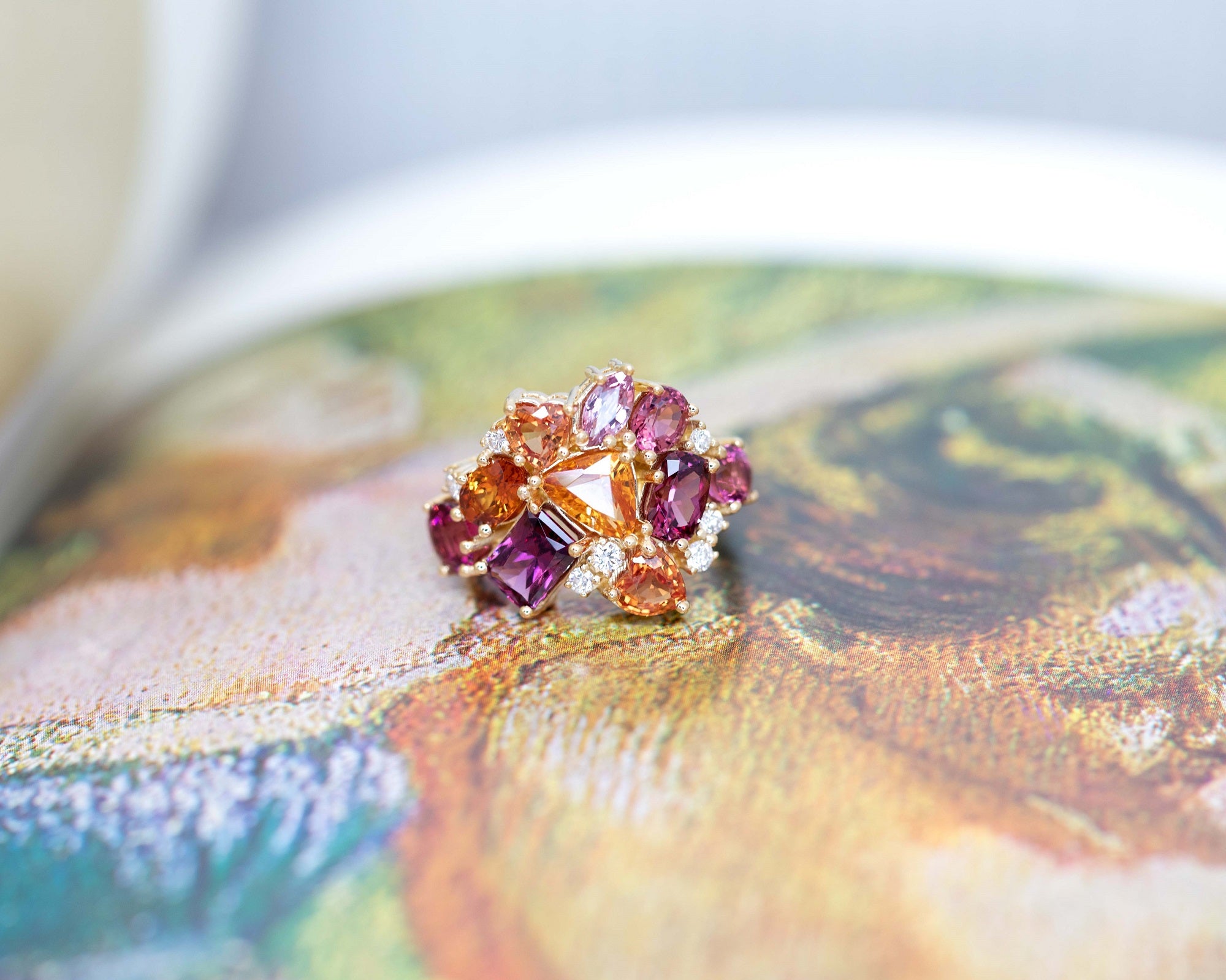 The Avalanche Lavish Lava Ring against a colored background, emphasizing the fiery brilliance of the gemstones and the warm yellow gold setting. Designed by Bena Jewelry in Montreal, Canada.