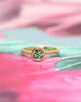 Elegant Bezel Set Demantoide Ring in yellow gold, featuring a natural green garnet, showcased on a soft pink and floral background. Made in Canada.