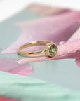 Yellow gold ring with a bezel-set green demantoid garnet by Bena Jewelry, shown on a rose-hued floral background. Minimalist bridal jewelry.