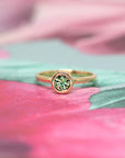 Bena Jewelry’s Bezel Set Demantoide Ring, a minimalist yellow gold ring with a green garnet, displayed on a delicate pink flower-themed background.