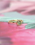 Bezel Set Demantoide Ring by Bena Jewelry, with a vibrant green garnet set in yellow gold, displayed on a soft pink floral background. Handmade in Montreal.