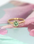 Green demantoid garnet engagement ring by Bena Jewelry, photographed against a romantic pink floral backdrop. Custom-made in Montreal.