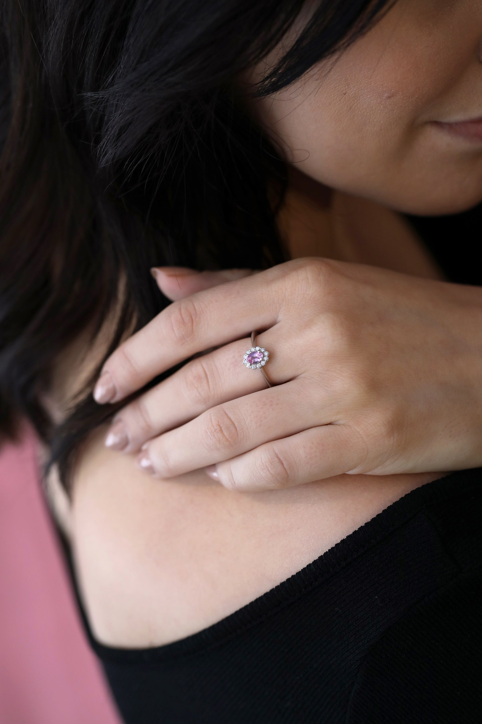 White gold engagement ring featuring an oval pink sapphire surrounded by diamonds, Diamond Halo Pink Sapphire Ring by Bena Jewelry, shown in a close-up on a woman’s hand.