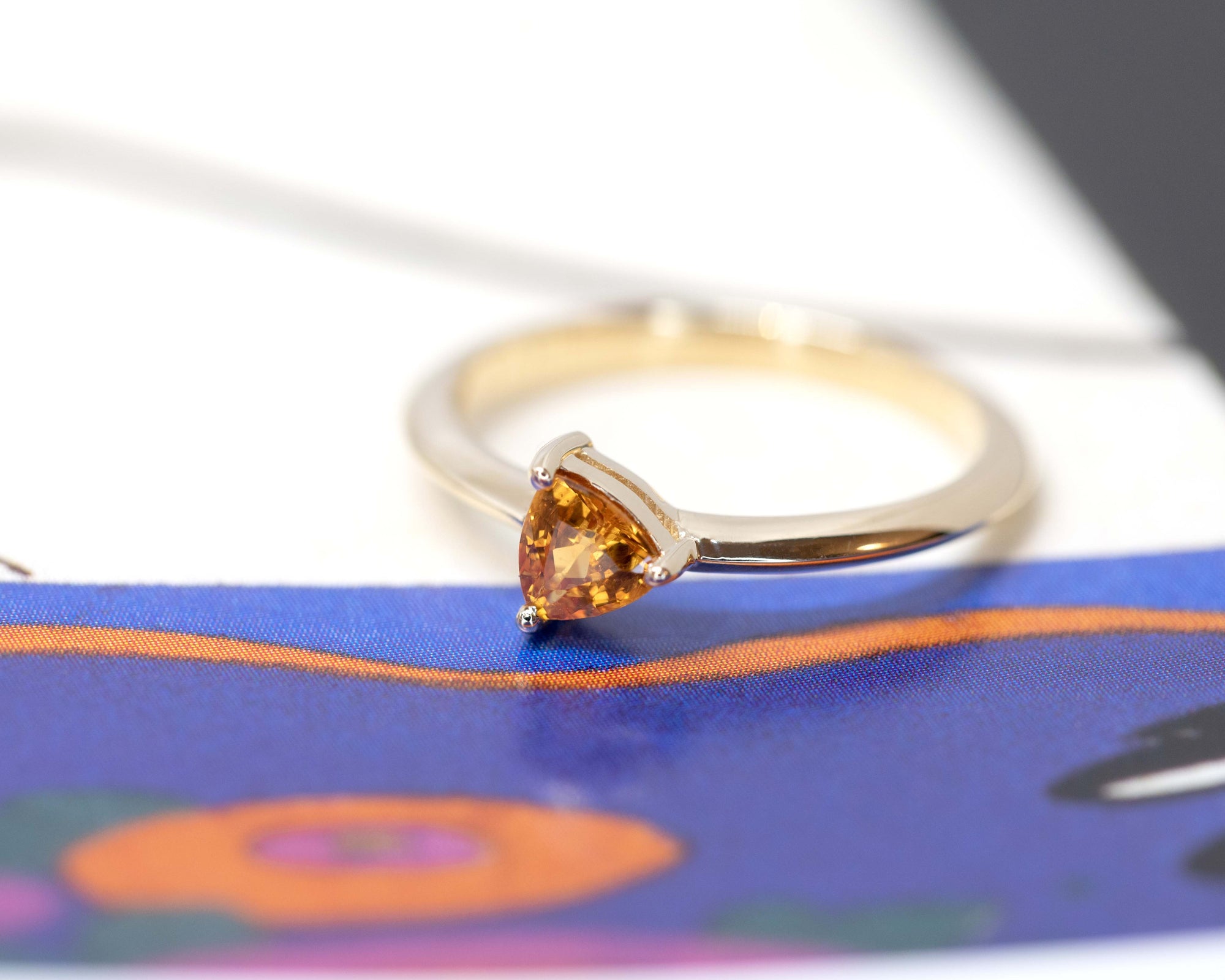 side view of trillion shape orange sapphire gemstone minimalist design custom made in montreal by the designer bena jewelry on a multi color background