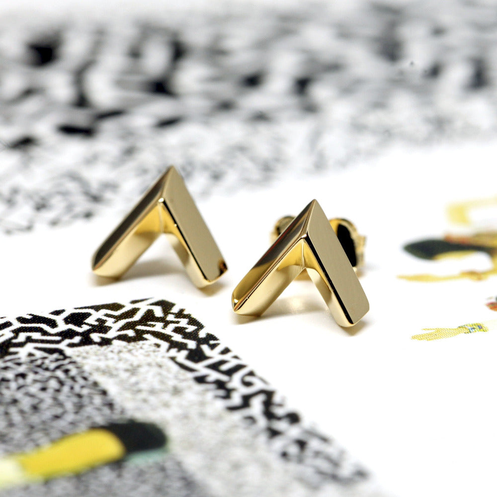 Yellow gold Arrow stud earrings displayed on a vibrant colored background. The edgy, minimalist, and unisex design highlights the modern craftsmanship of Bena Jewelry, made in Montreal.