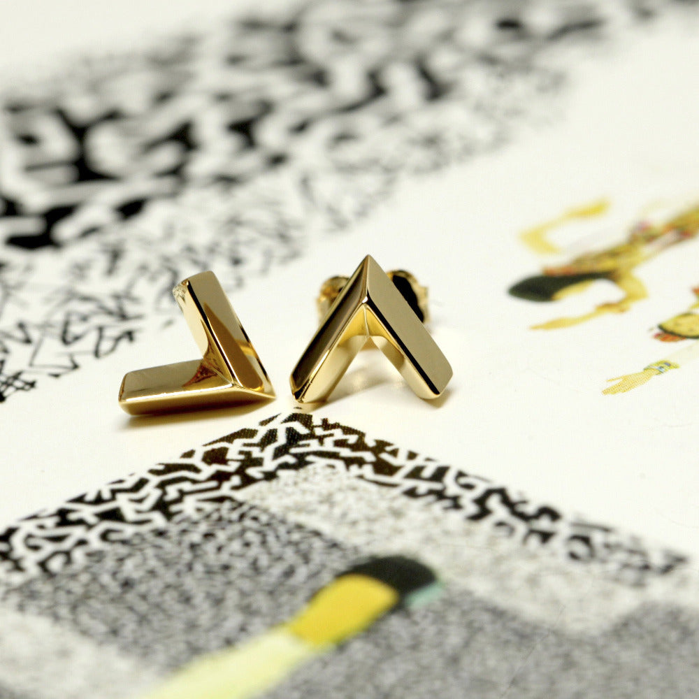 Arrow studs in yellow gold showcased against a bold, colorful background. A sleek and contemporary unisex design by Bena Jewelry, crafted in Montreal, Canada.