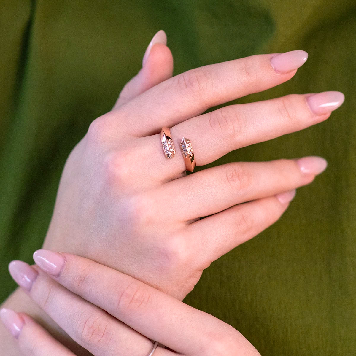 A close-up of hands wearing an elegant open rose gold ring featuring diamond accents on both ends. The modern, minimalist design complements the soft pink nails and green outfit, creating a refined and contemporary look.