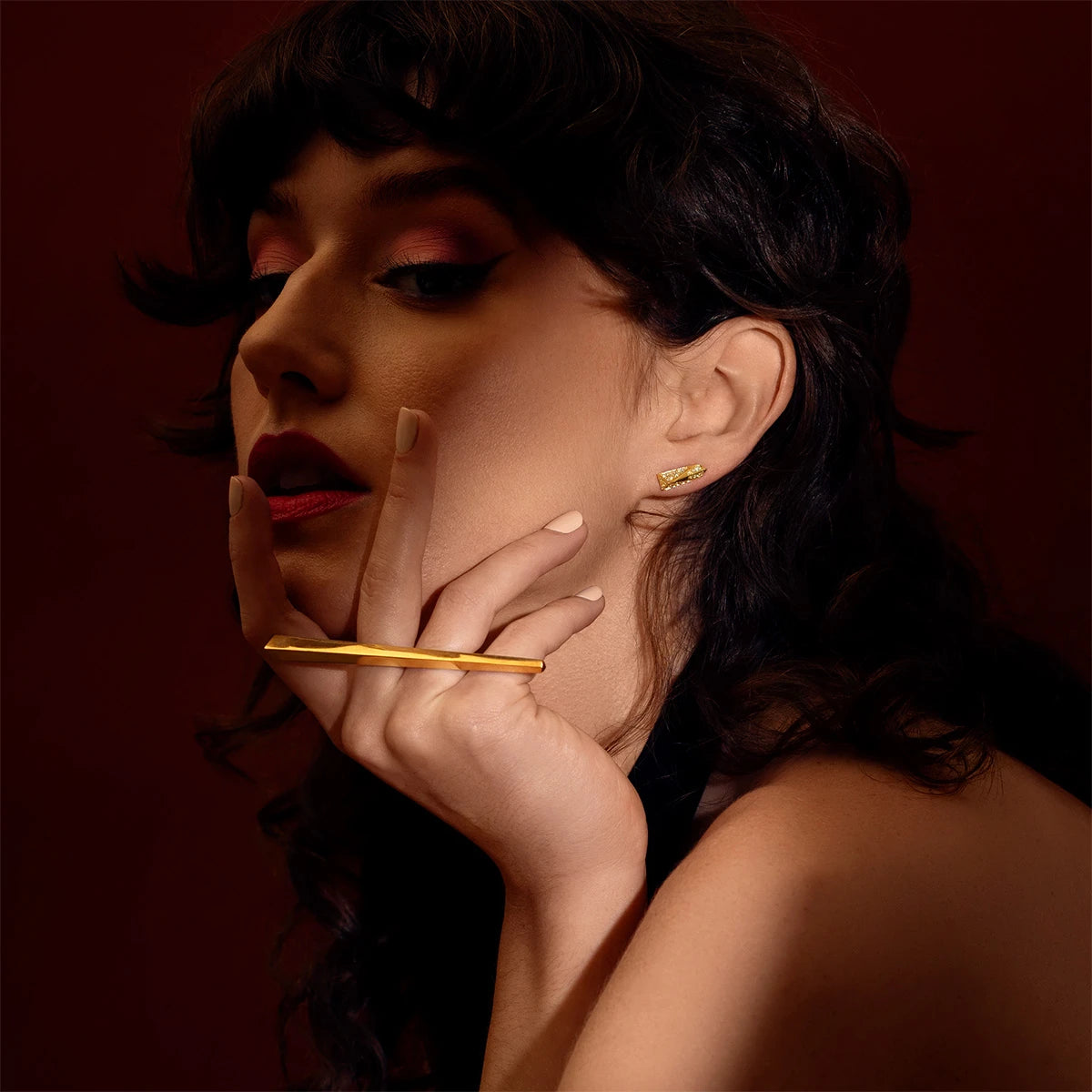 A beautiful young woman with dark brown curly hair is wearing 2 pieces of unisex fashion modern gold jewellery. On her ear, she wears a bold yellow gold diamond stud that shows a lot of volumes, and on her hand, a long and slick double finger ring in yellow gold. Both high end jewelry pieces were designed by Bena Jewelry.