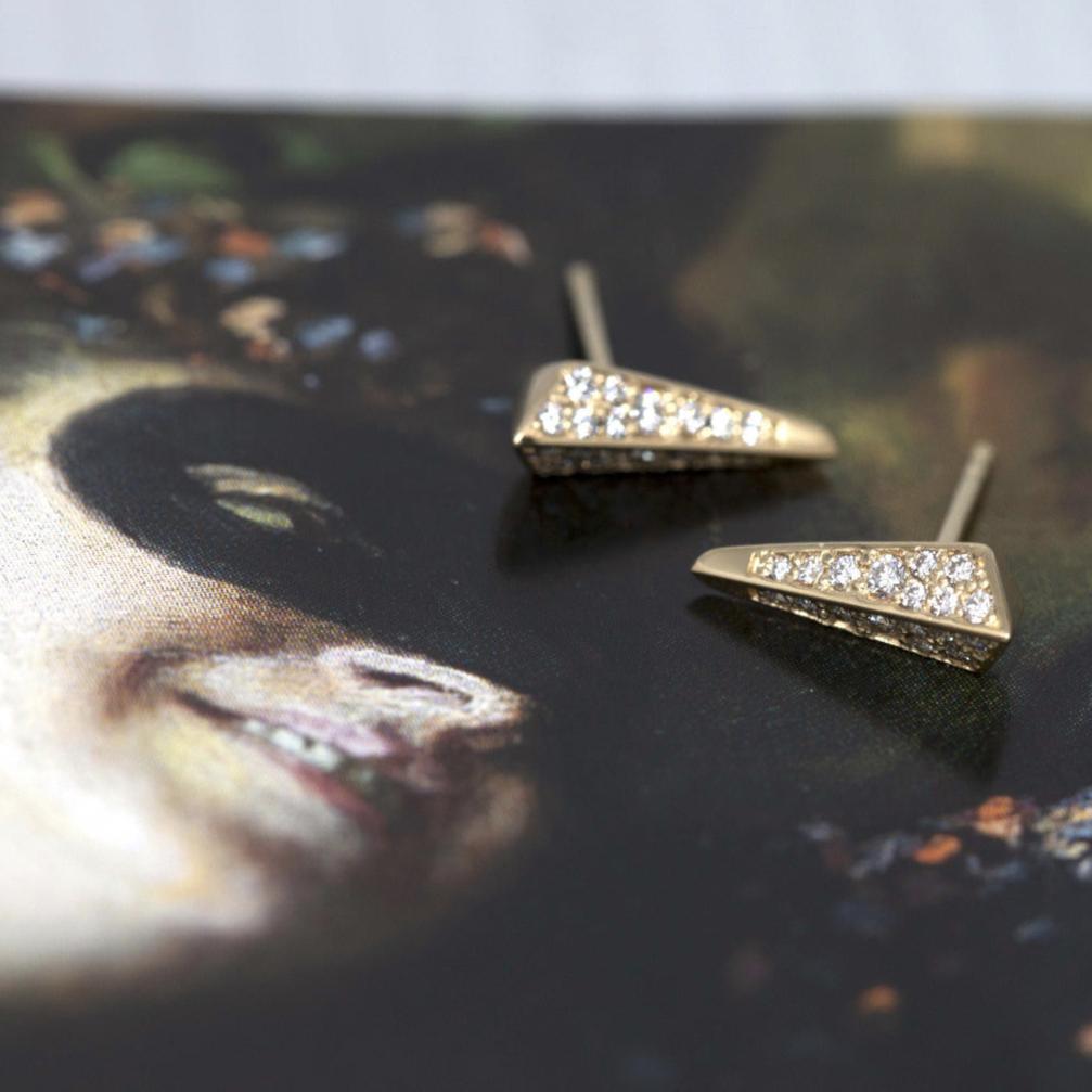 A pair of gold buckles with diamond pavé is photographed on a carnival image of a woman wearing a mask. The earrings are unisex and have a modern, elegant and chic style. Created by independent designer Bena Jewelry.